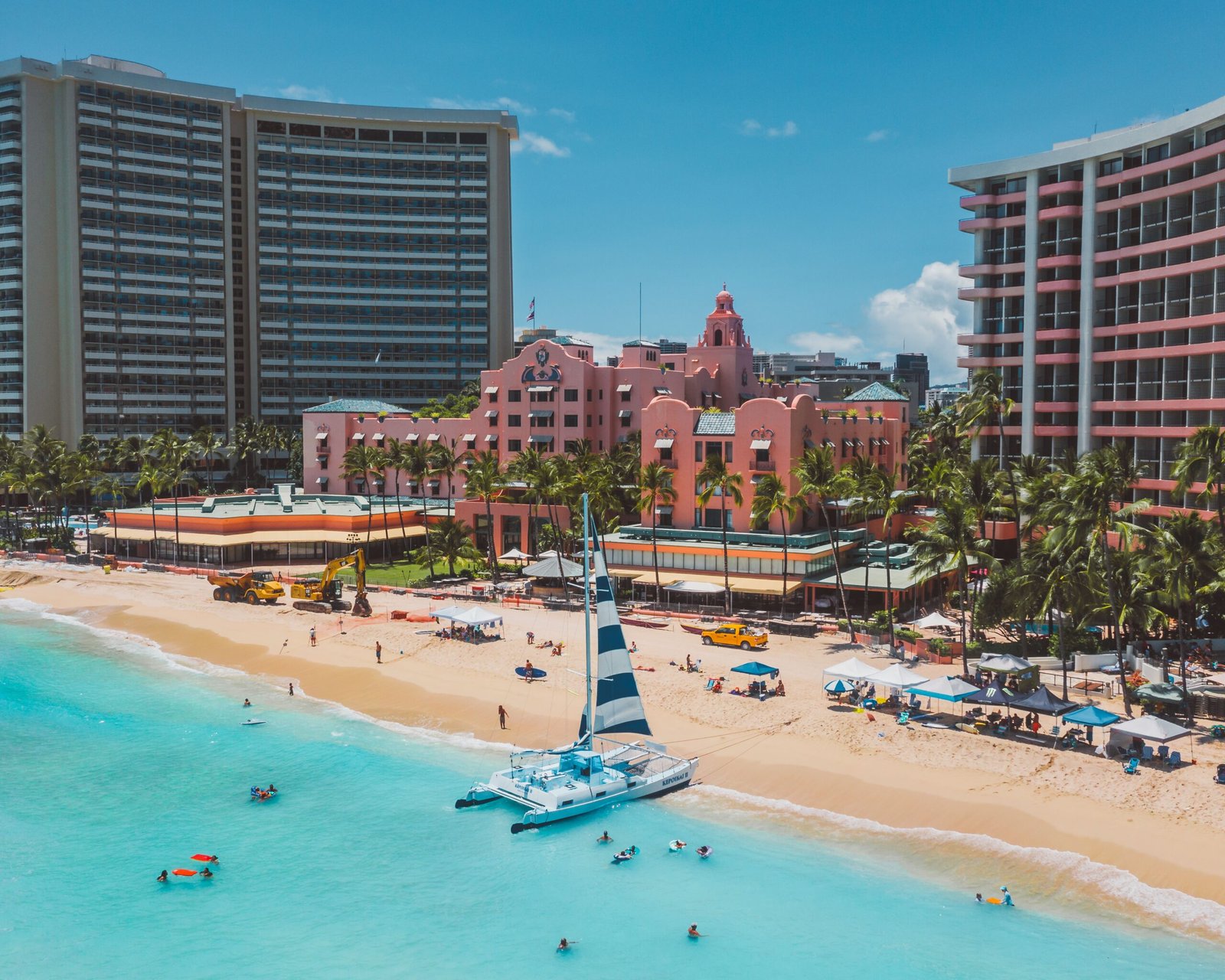 Aerial view of a vibrant beach resort with pink buildings, palm trees, and turquoise waters, highlighting leisure activities.